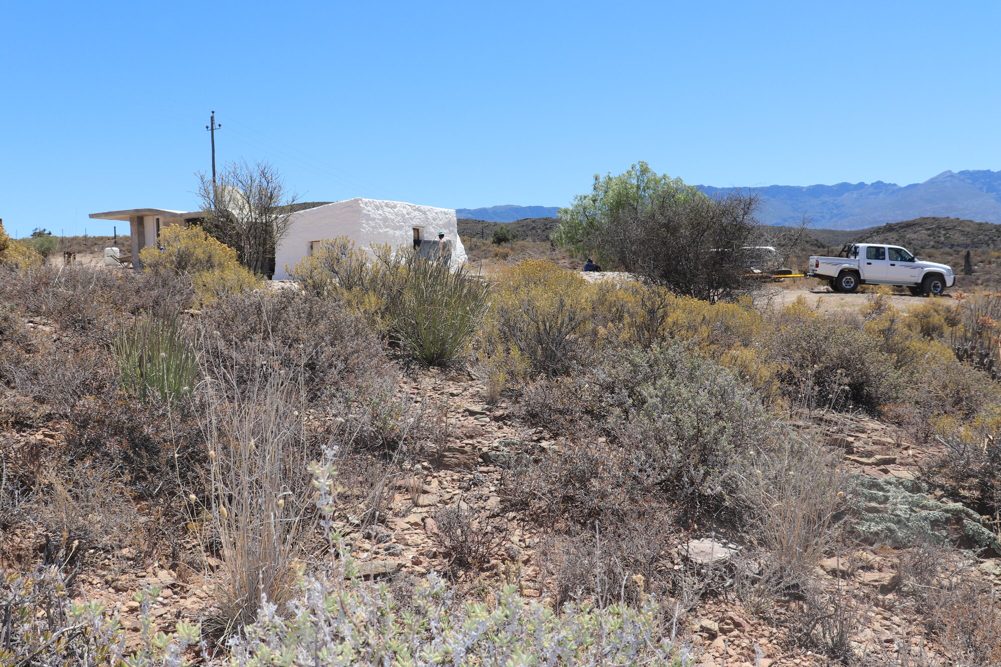 Sheep Herder's Cottage in its Klein Karoo setting — a small lime-washed building with a freeform concrete shell roof, open veld and mountains beyond